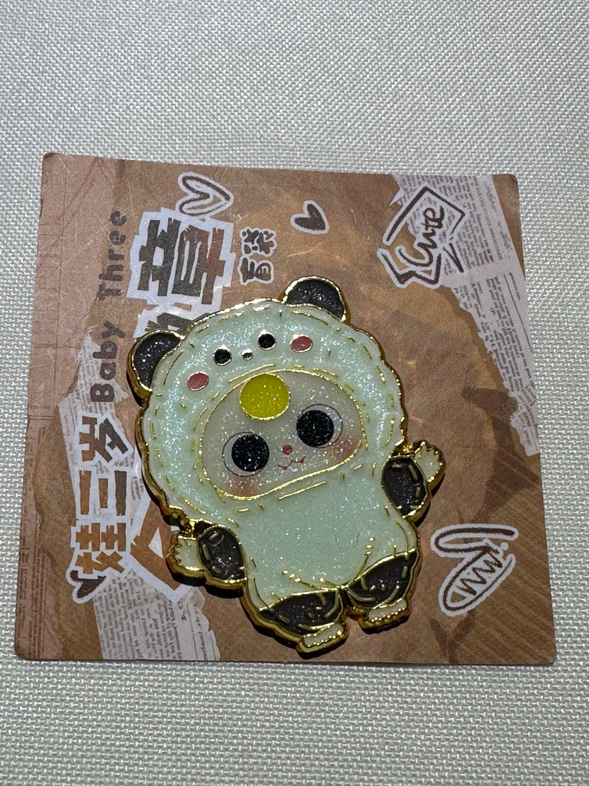 Baby Three Panda enamel pin with black ears and paws and yellow forehead detail on backing card