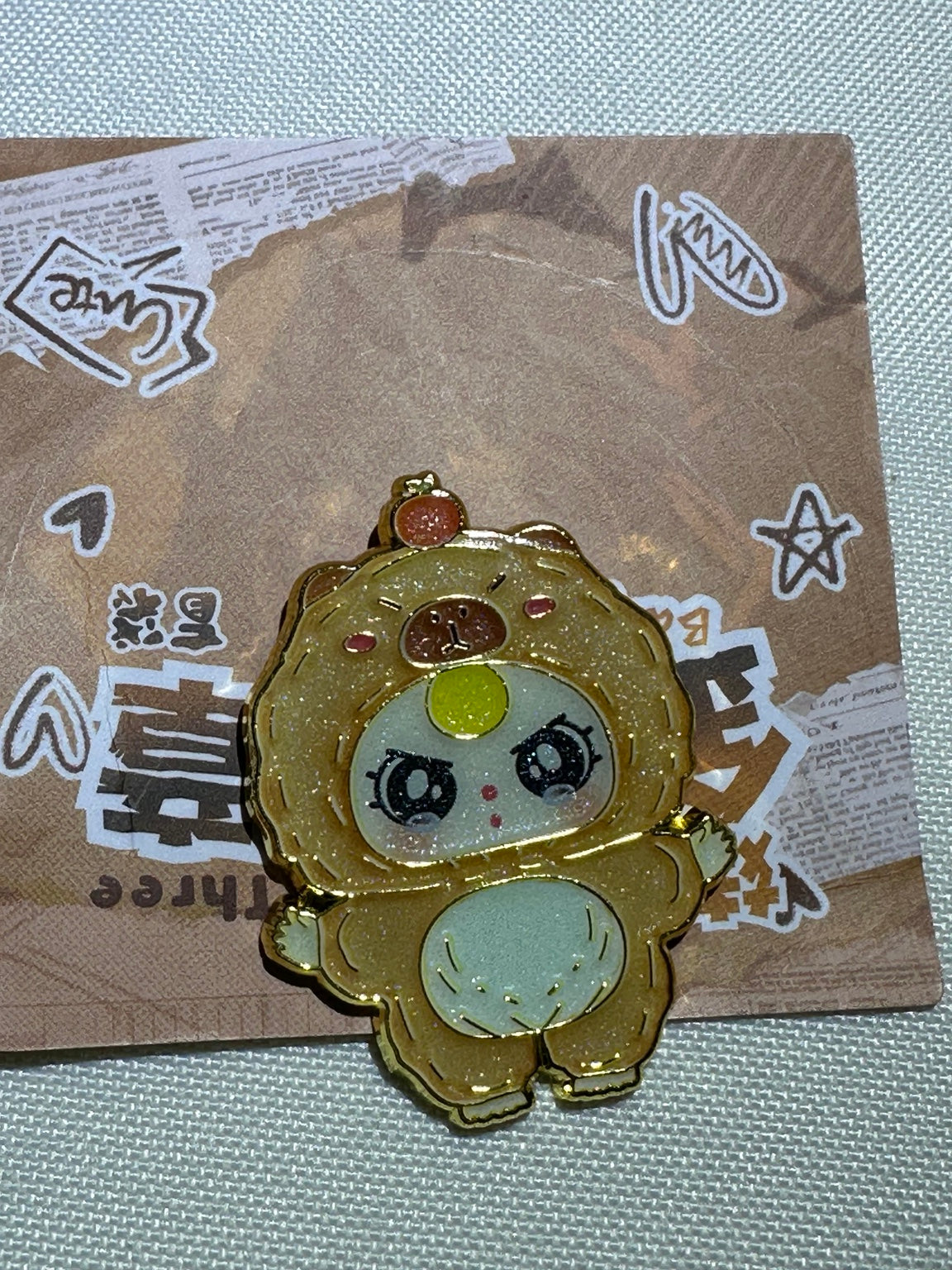 Baby Three Capybara enamel pin in tan with a pale belly and yellow forehead accent on backing card