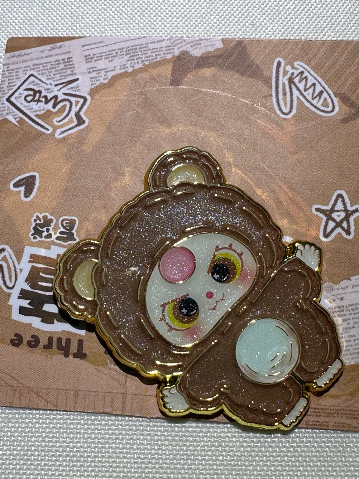 Baby Three Brown Bear enamel pin with pale belly, pink forehead accent, and gold-colored outlines on backing card