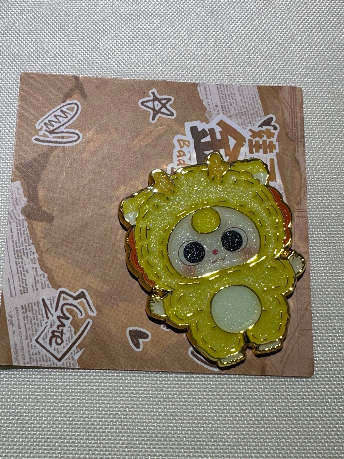 Baby Three Lion enamel pin in yellow with a pale belly and gold-colored outlines on backing card